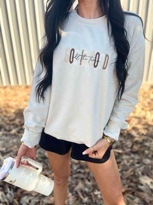 A person wearing a light-colored, embroidered sweatshirt with the text 'GOD IS GOOD' on the front, standing outdoors.