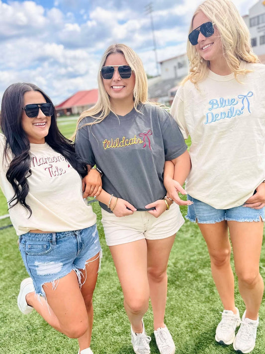 Three women standing on a grassy field wearing casual outfits with visible mascot school text on their shirts.