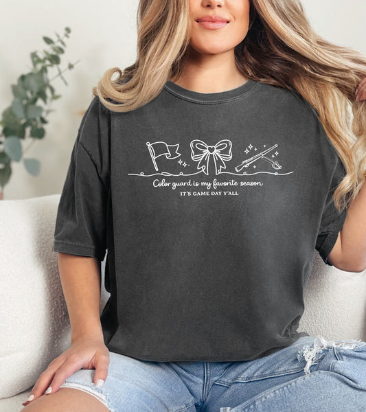 Person wearing a dark gray t-shirt with a "Color Guard is my favorite season" graphic design and text, sitting on a couch.